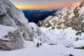 Zwei Bergwanderer beim eisigen Kletteraufstieg. Im Hintergrund der Sonnenaufgang im Bergpanorama.
